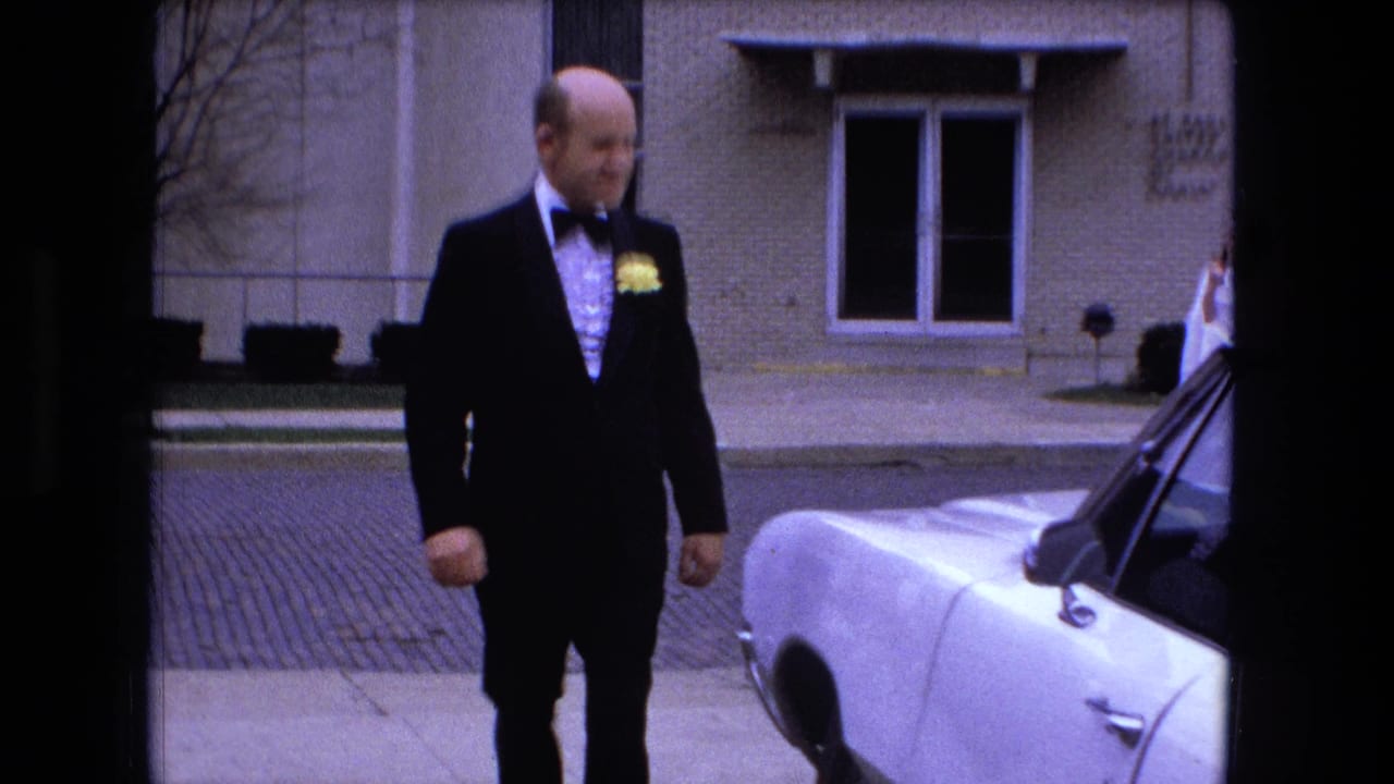Tuxedoed man stands closer to a white car on a street outside a brick building.