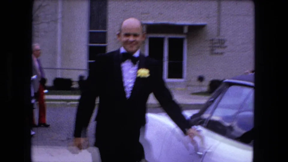 A smiling tuxedoed man reaches for the door of a white car in Fort Wayne, Indiana, captured on 1973 8mm home-movie film.