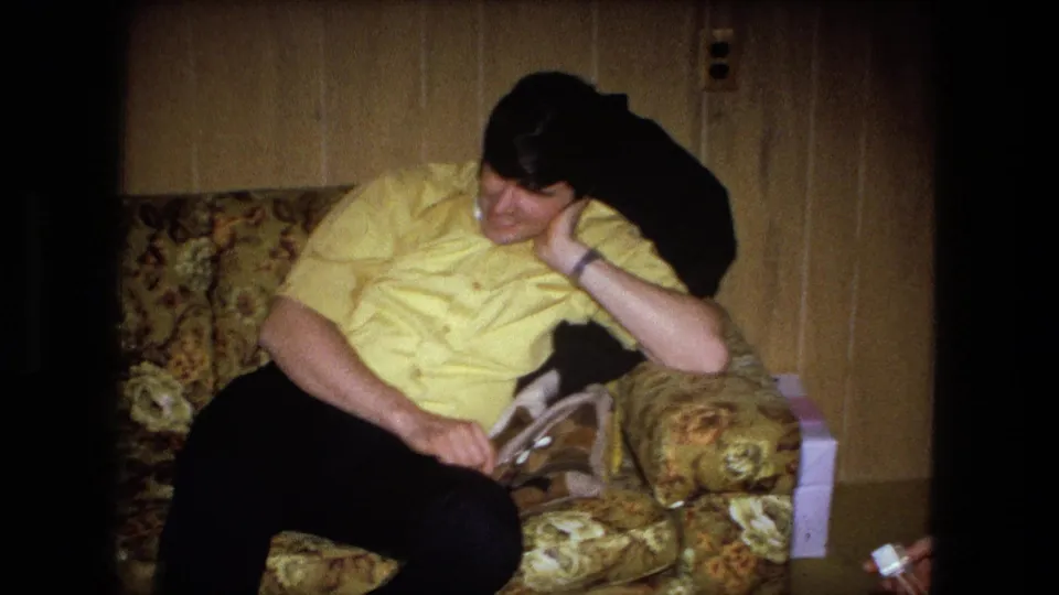 A man in a yellow shirt relaxes on a floral-patterned sofa inside a wood-paneled room in the same 1973 home-movie clip.