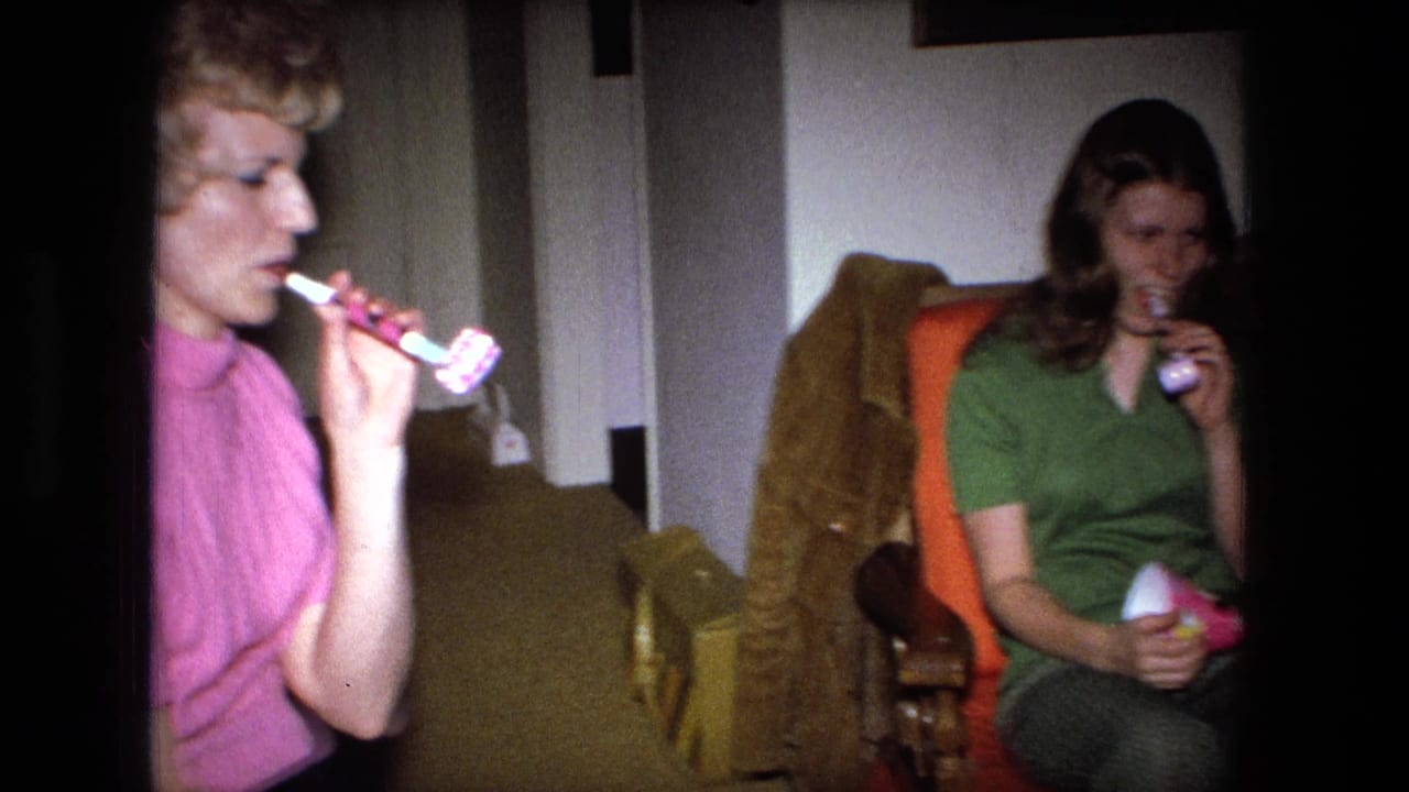 Two women in a living room blow party horns while seated near upholstered chairs.