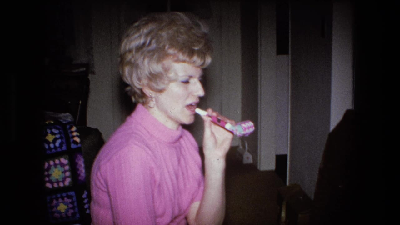 A blonde woman in a pink ribbed sweater blows a colorful party horn in a Fort Wayne home interior, 1973.