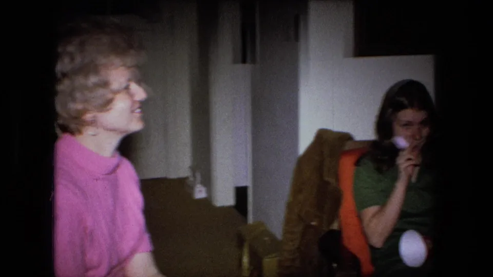 Two adult women sit in chairs with party horns in a dim 1973 Fort Wayne living-room party scene.