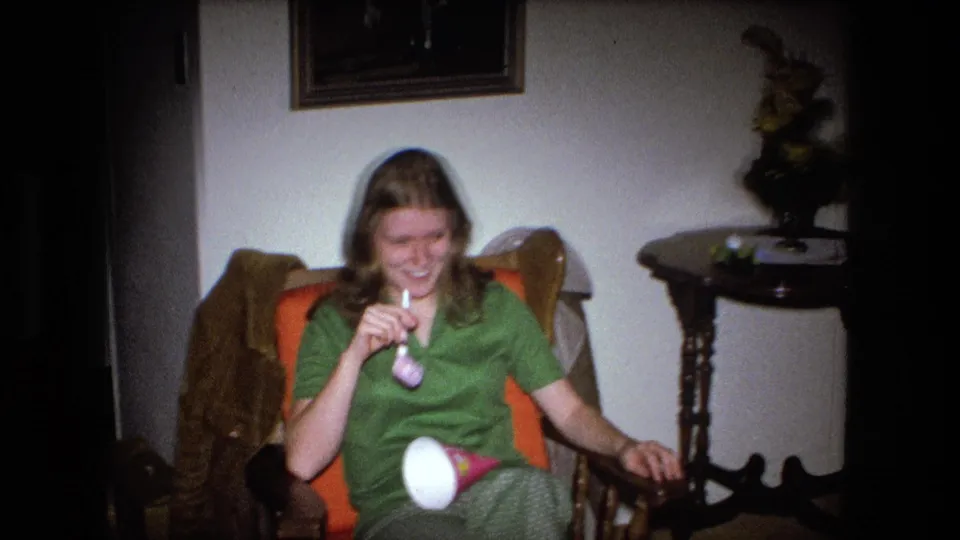 A woman in a green top laughs with a party horn while seated near a side table in a 1973 Fort Wayne home.