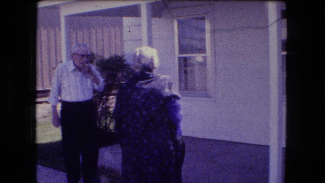 Older man and older woman standing by a porch and window outside a house.