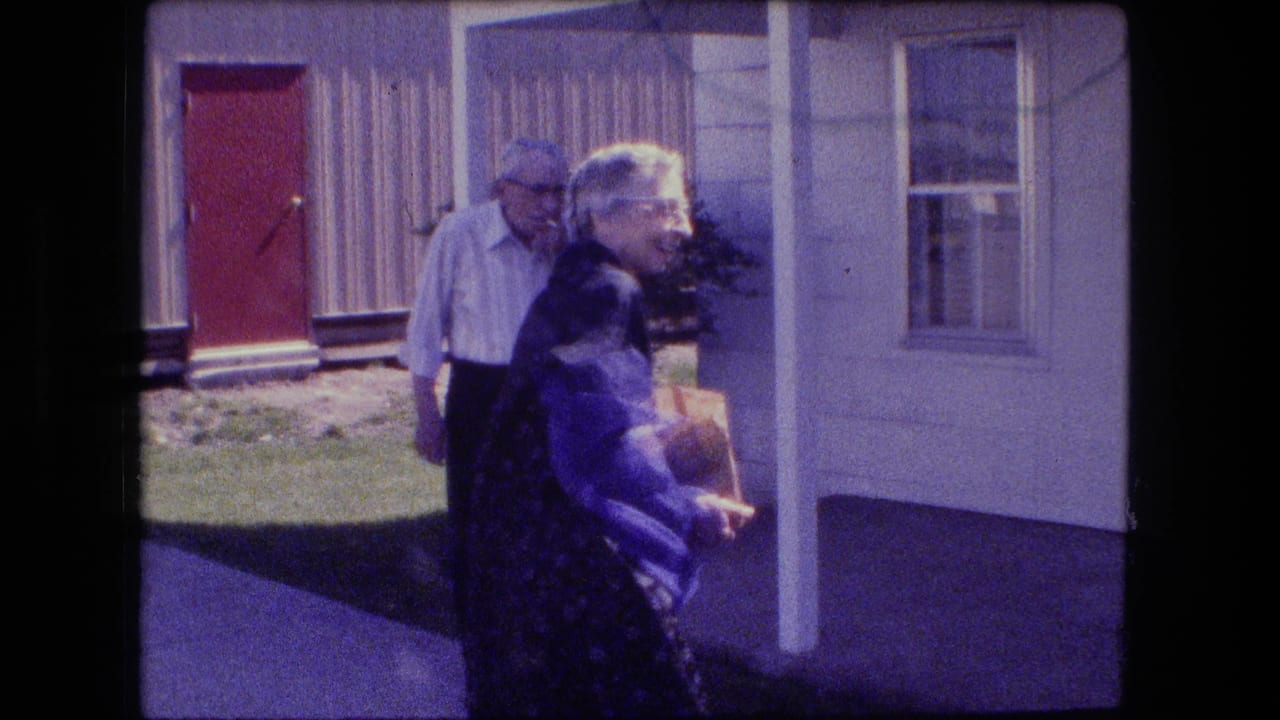 Older woman carrying a bag walks past an older man near a red front door.