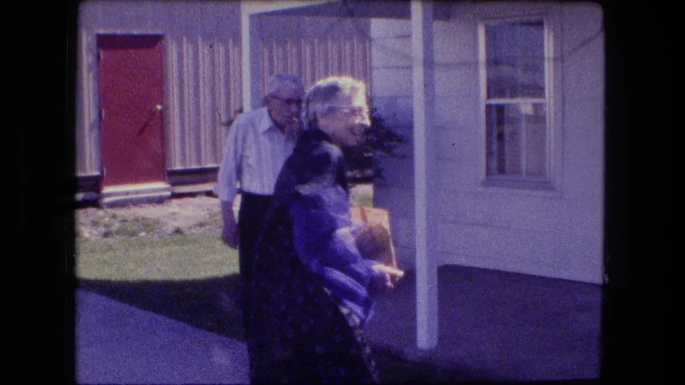 In 1973 Fort Wayne, an older woman with a bag moves across a porch area while an older man stands near a red front door.