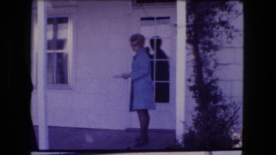 A woman in a light coat stands at the front door of a Fort Wayne residence in 1973 home-movie footage, framed by a porch post and shrub.