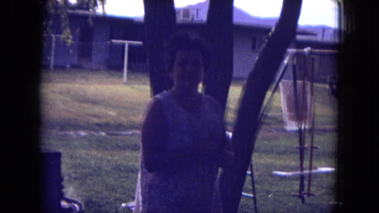 1973 8mm frame from New Mexico showing an adult woman in shade near trees, with a backyard swing set and house in the background.