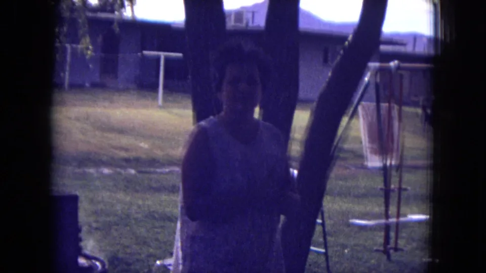 1973 8mm frame from New Mexico showing an adult woman in shade near trees, with a backyard swing set and house in the background.