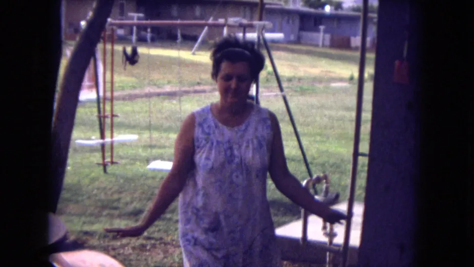 1973 New Mexico home-movie frame of a woman in a light patterned dress walking through a backyard near a swing set.