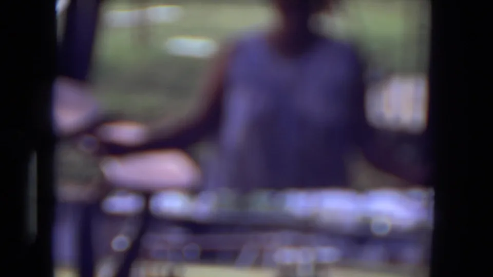 Soft-focus 1973 home-movie frame with the woman blurred near backyard furniture or equipment and green lawn in the background.