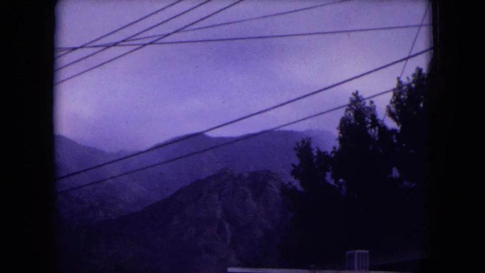 Purple-shifted 8mm frame of New Mexico mountain slopes, silhouetted trees, and diagonal utility lines under a hazy sky.