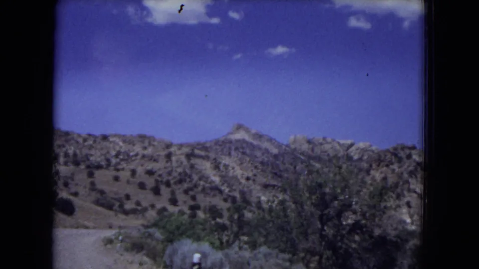 A moving road-view frame shows dry hills, sparse trees, and scrub in 1973 Gila National Forest footage.