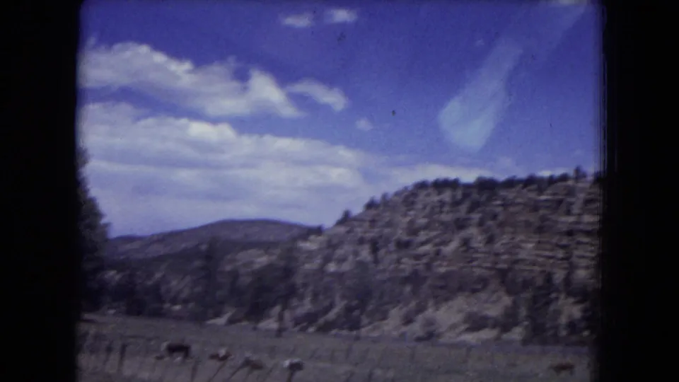 Gila National Forest landscape with layered rock slopes, scattered pine trees, open meadow, and bright clouds in the sky.
