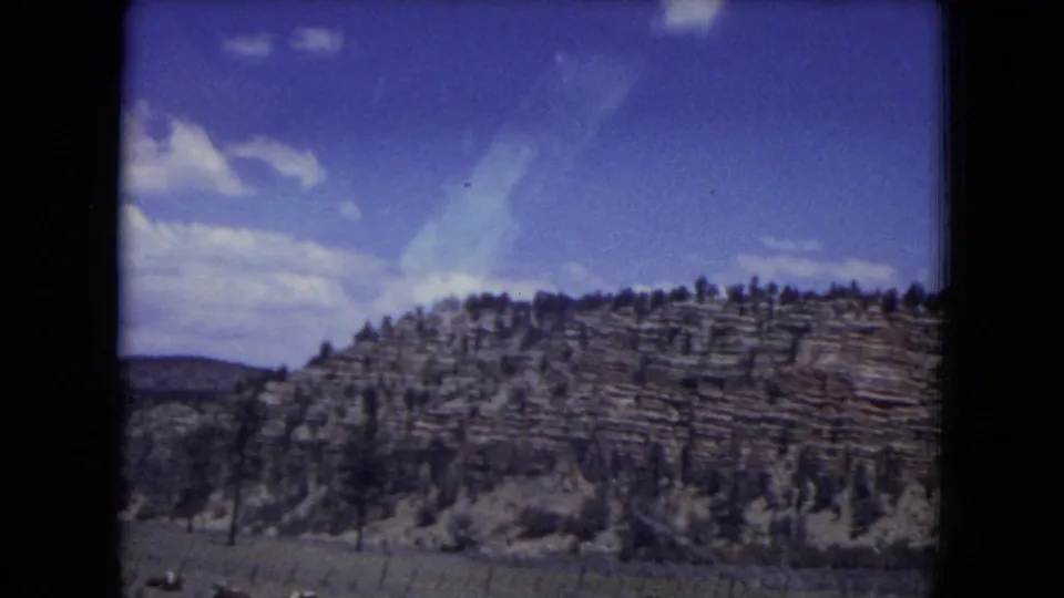 1970s 8mm view of a rocky Gila National Forest hillside with trees, meadow foreground, and small clouds overhead.