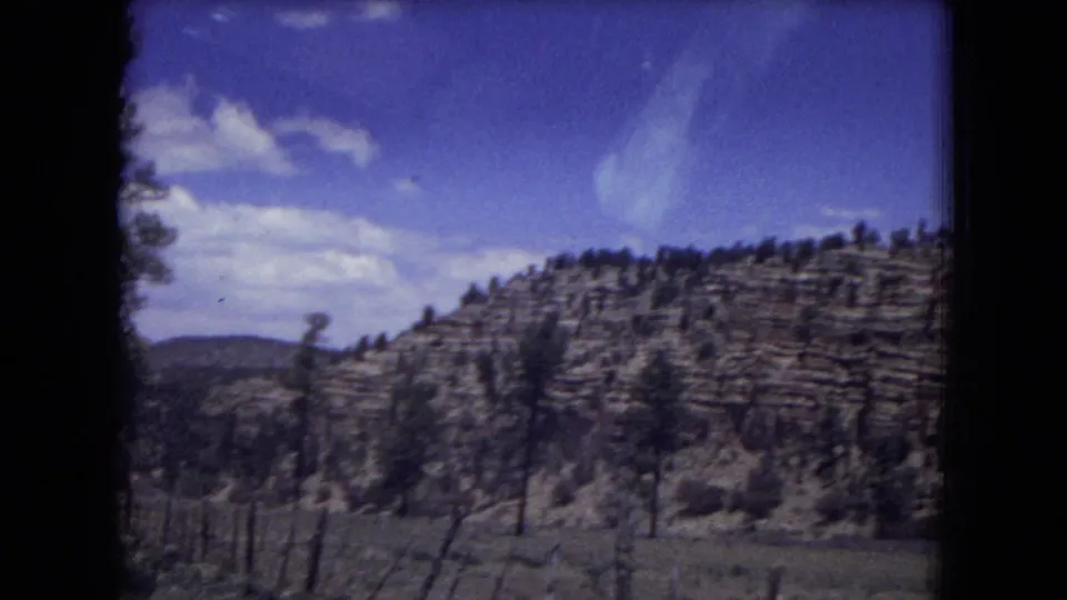 Gila National Forest view with a layered rock formation, sparse trees, meadow grasses, and blue sky on 8mm film.