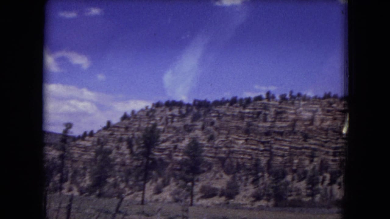 Layered rocky hillside with scattered pine trees under blue sky, seen through a dark window frame.