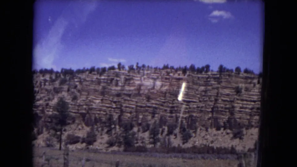 Gila National Forest hillside with horizontal rock layers and pine trees, captured on 1973 8mm film with visible flare.