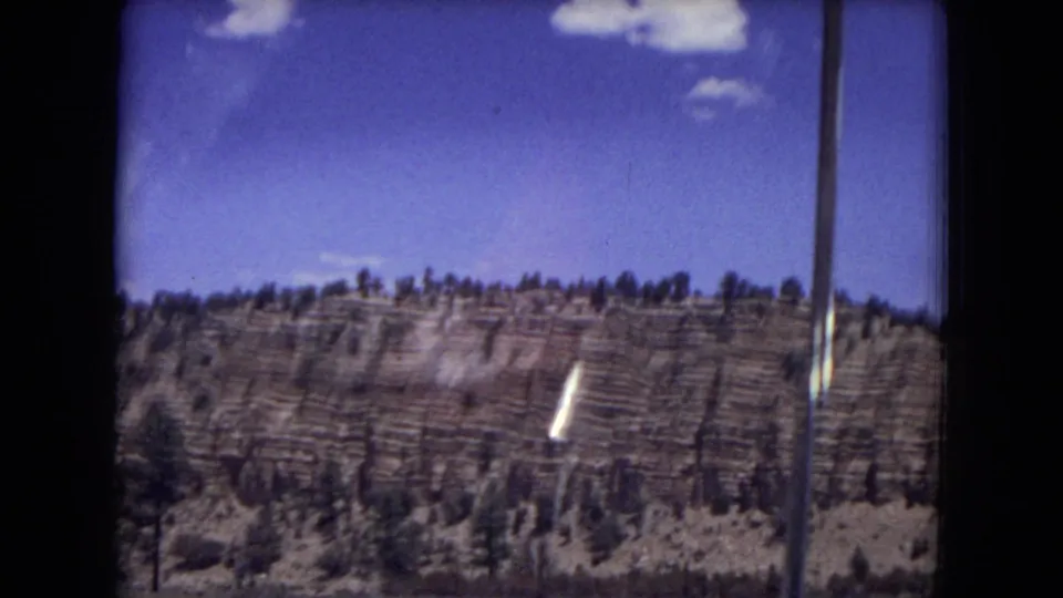 1973 home-movie frame of Gila National Forest terrain, showing layered rock, pines, and a vertical foreground post.