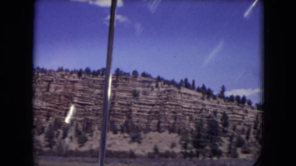 Gila National Forest mountain slope in a 1973 8mm frame, with pine trees, horizontal rock bands, and window framing.