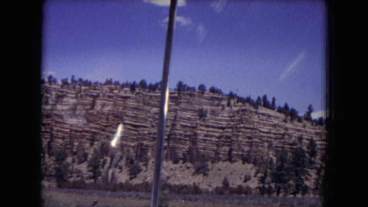 Layered cliff face with scattered pines below a blue sky, seen through dark 8mm frame edges.