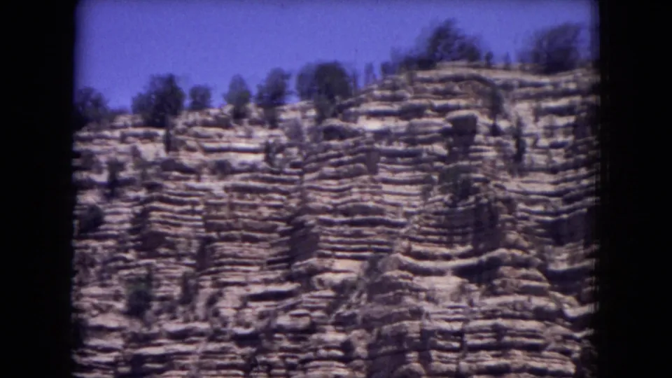 Close 8mm frame of banded rock formations in Gila National Forest, with small trees on the ridge and visible film softness from motion.