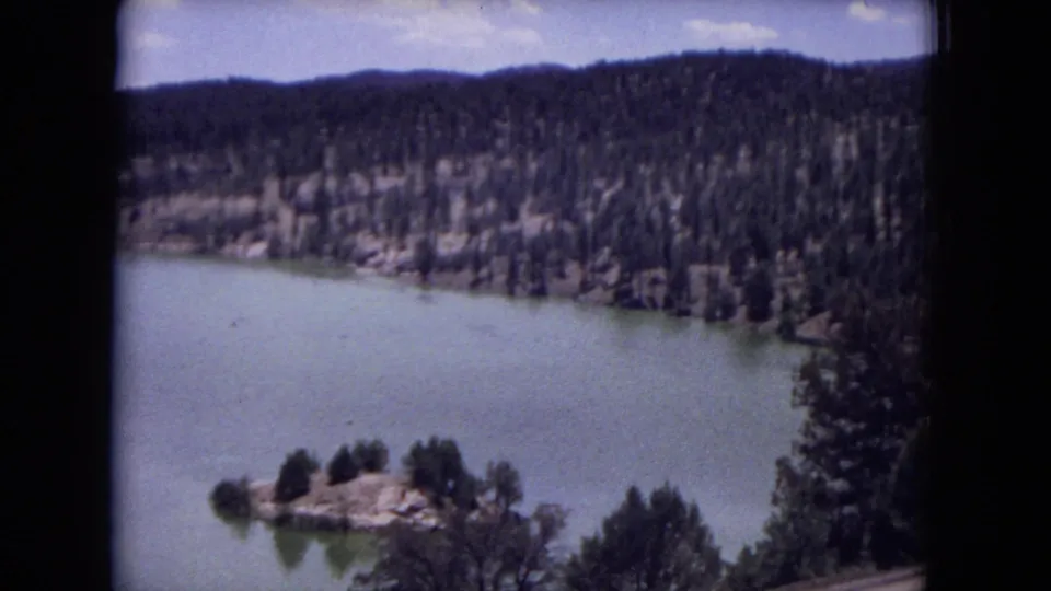 Gila National Forest lake view from 1973 8mm footage, with pale water, wooded hills, shoreline trees, and a hazy blue sky.