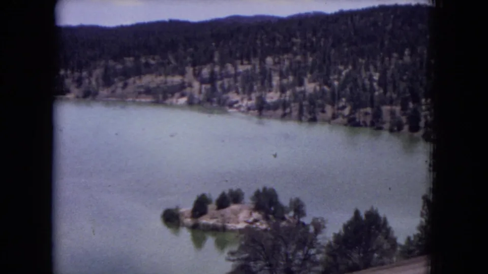 1973 8mm view in Gila National Forest, New Mexico, showing calm water, a wooded island, and pine-covered hills from above.