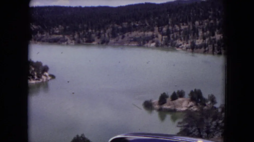 Gila National Forest lake scene on 8mm film, with small tree-covered islands, pale water, wooded hills, and frame-edge obstruction.