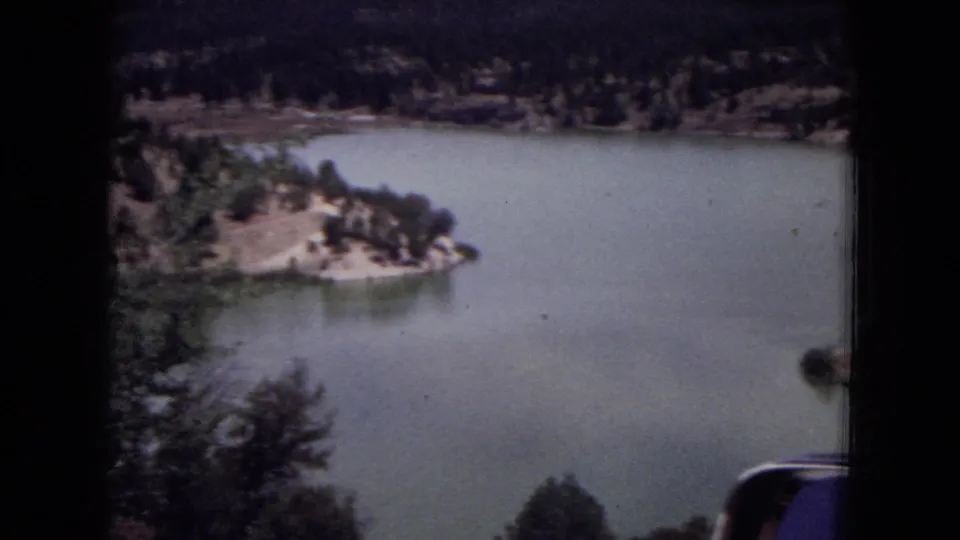 1973 home-movie frame of a New Mexico forest lake, showing a wooded peninsula, still water, foreground branches, and distant shoreline.