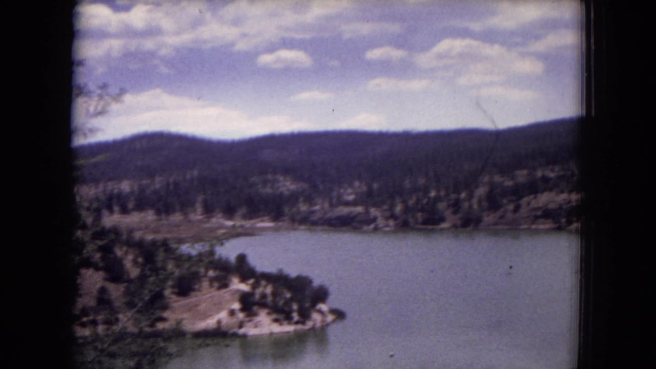 Calm lake below forested hills under a partly cloudy sky, framed by dark film borders.