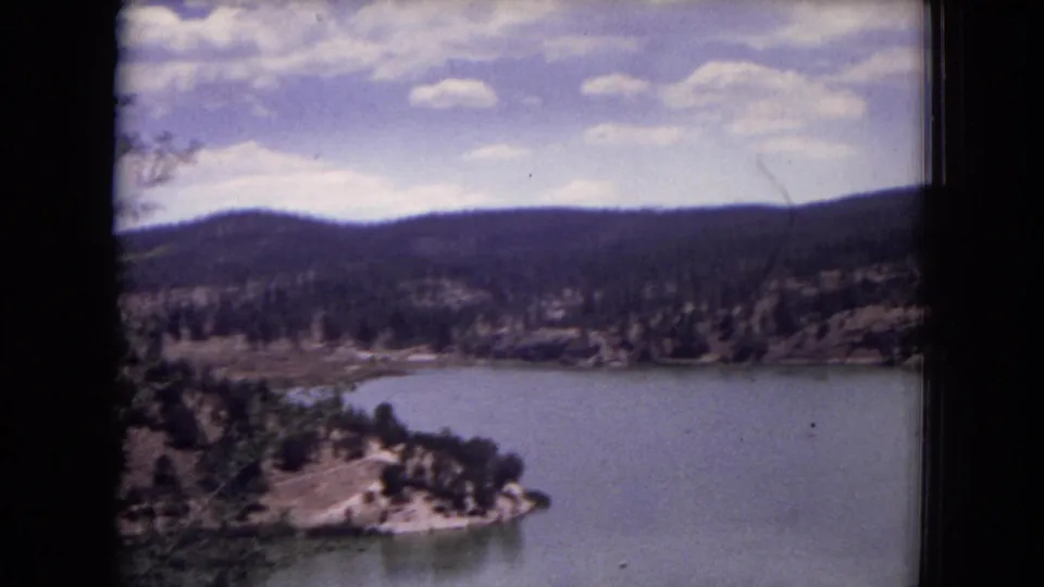 1973 8mm home-movie view of a lake and pine-covered hills in Gila National Forest, New Mexico, seen through a dark border.