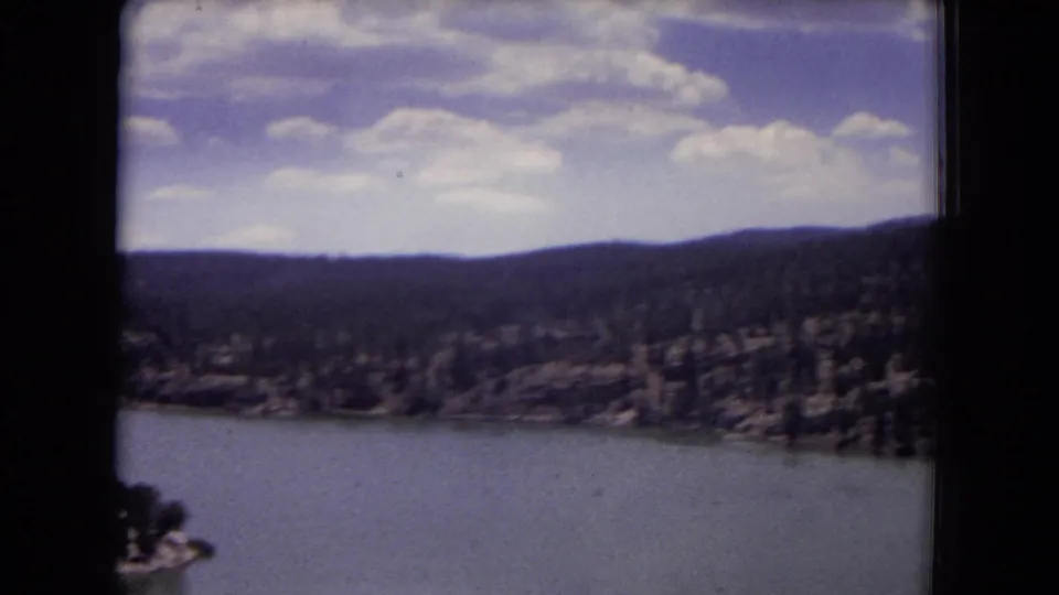 Soft 8mm view across a lake toward wooded hills in the Gila National Forest area, with cloudy sky and dark frame edges.