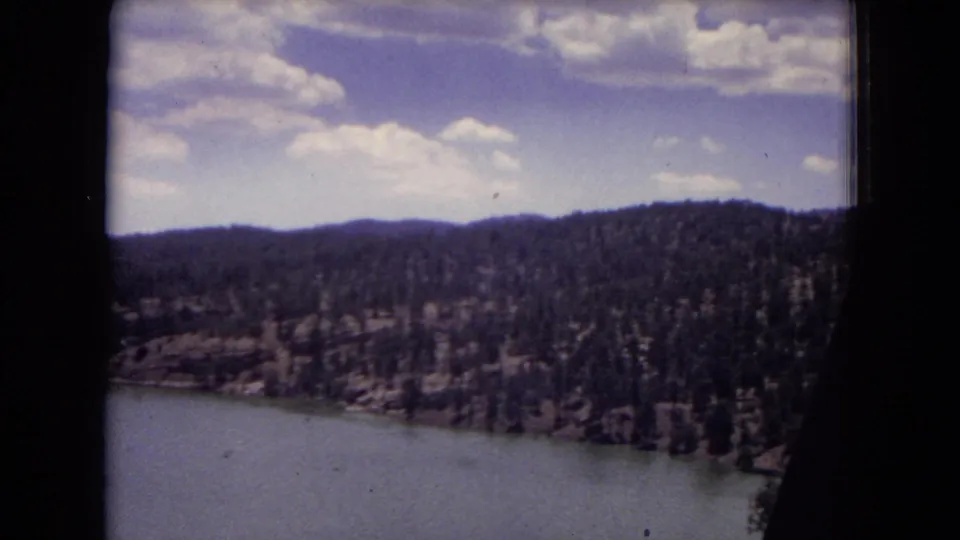 1973 8mm frame showing lake water below a forested slope, likely in Gila National Forest, New Mexico, during a slow scenic pan.