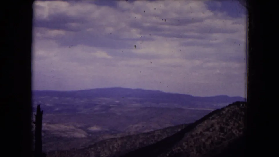 Wide 1973 home-movie view across Gila National Forest hills and distant ridges, with a cloudy sky and a dark slope at the lower right.