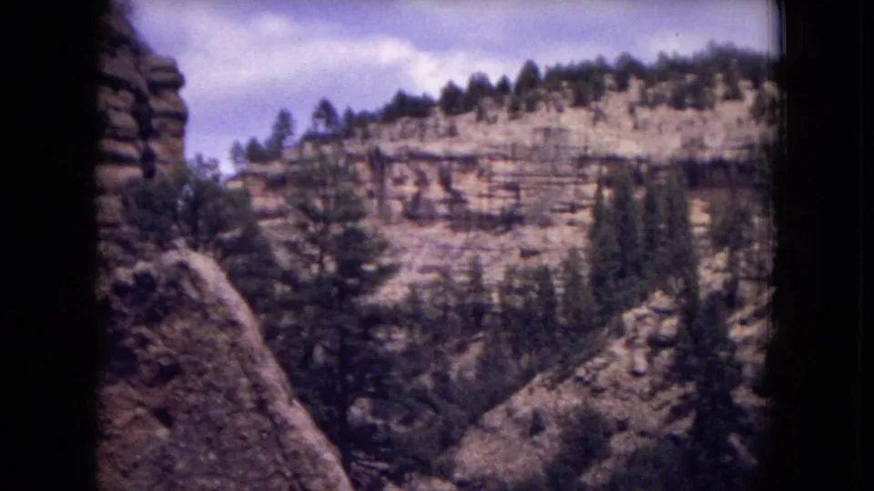1973 8mm frame showing layered cliff bands and pine trees across a canyon in Gila National Forest, New Mexico.