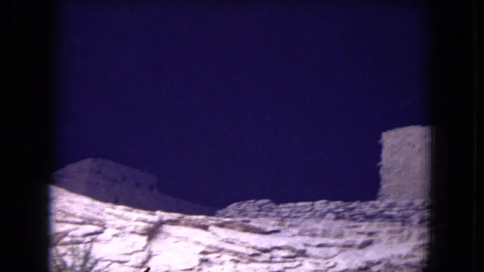 1973 8mm frame from Gila National Forest showing weathered stone ruin walls on pale rock beneath a deep blue sky.
