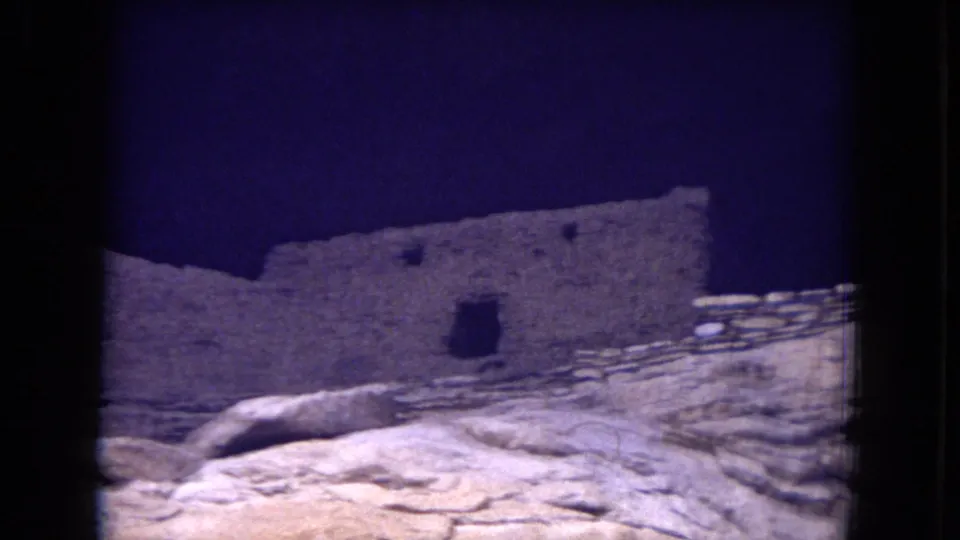 1973 8mm frame of weathered stone ruins in Gila National Forest, New Mexico, seen across pale rocky ground under a deep blue sky.