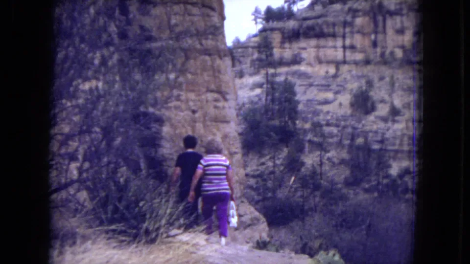 Two visitors seen from behind walk a narrow path below sandstone cliffs and scrub vegetation in the 1973 Gila National Forest footage.