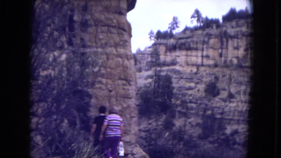 Visitors continue down a narrow path below tall rocky cliffs, with scrub plants and layered sandstone visible in this 1973 New Mexico home-movie frame.