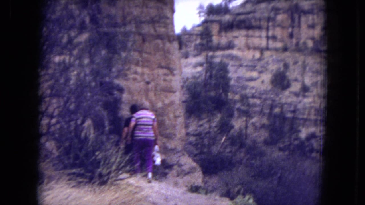 Woman walking along a narrow canyon trail beside scrub and sandstone cliffs.