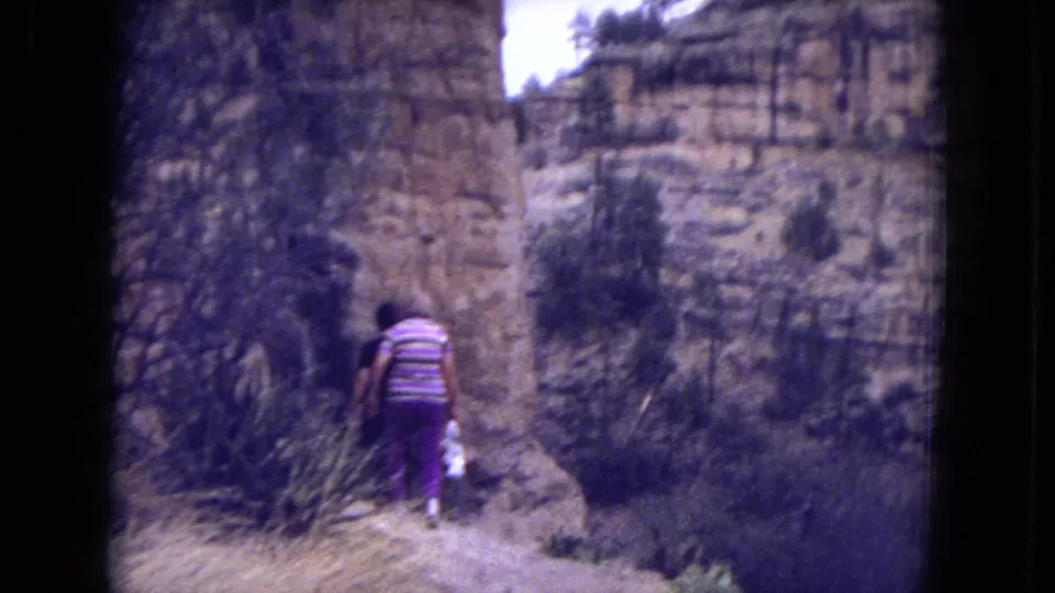 1973 8mm frame of a woman walking away on a rugged trail in Gila National Forest, with brush and sandstone canyon walls around her.