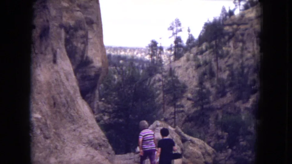 Rear view of two women hiking through rocky Gila National Forest terrain, with canyon walls and scattered pines in the distance.