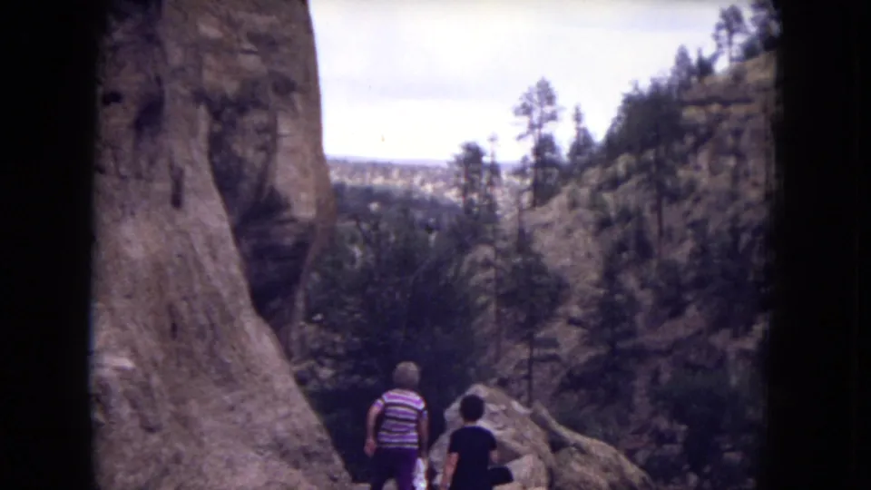 Wide 8mm view of two women walking through a rocky Gila National Forest canyon overlook with pine trees and distant terrain.