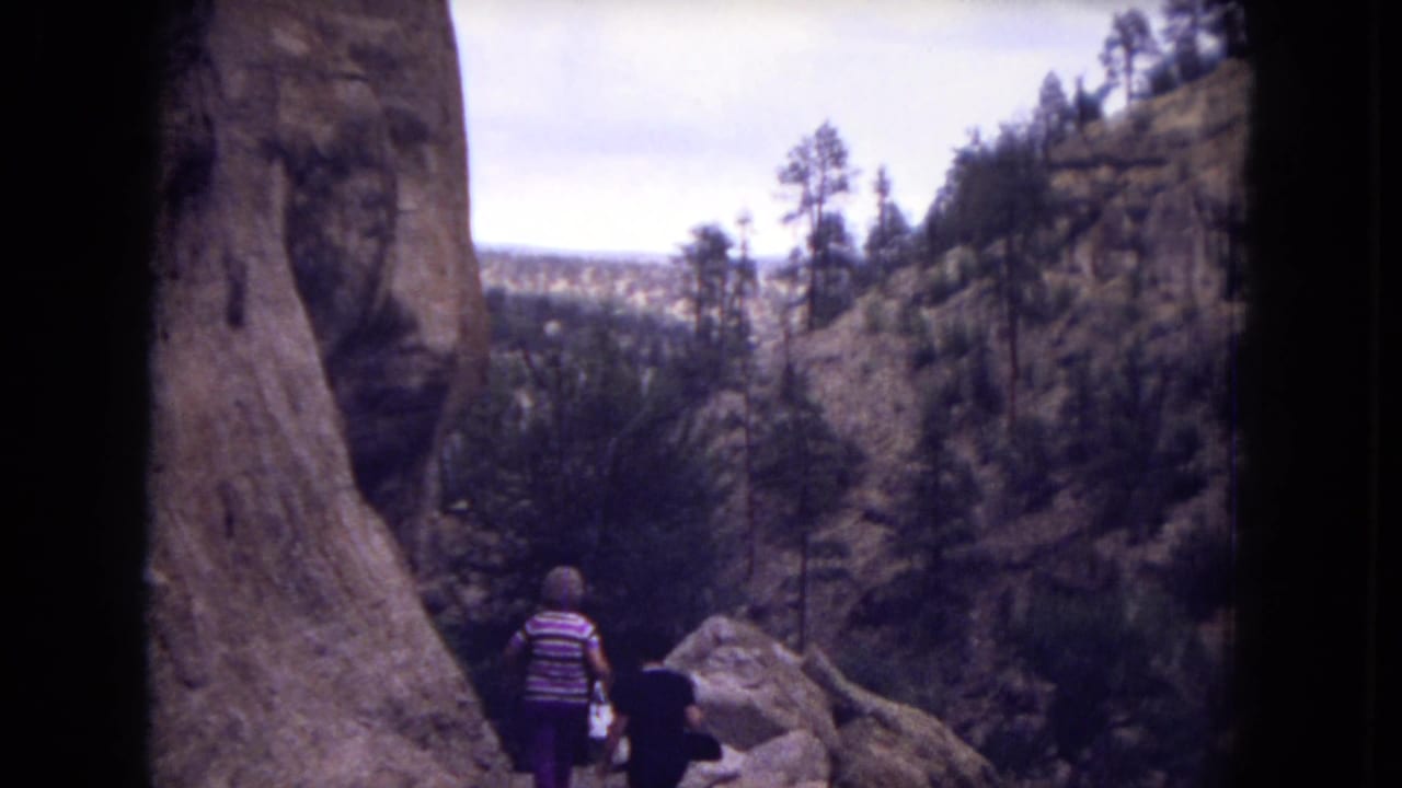 Two hikers walk along a rocky trail beside a tall canyon wall.