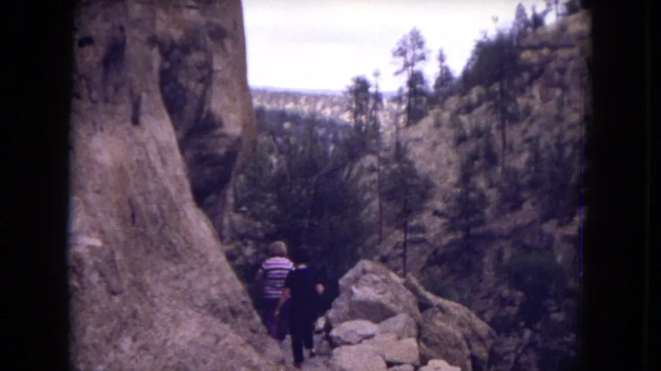 Two hikers walk away from the camera on a rugged New Mexico mountain trail, with rock formations and sparse pine trees around them.