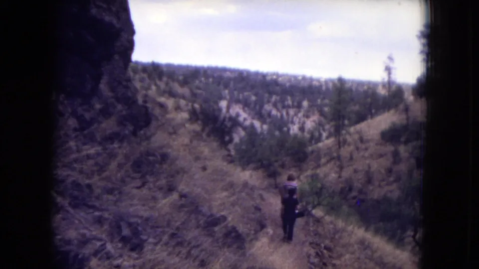 A distant hiker on a narrow dirt trail overlooks dry, pine-dotted canyon hills in Gila National Forest, New Mexico.