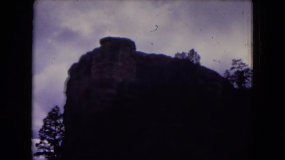 1973 Gila National Forest frame with a shadowed rock cliff, nearby trees, and a muted overcast sky.