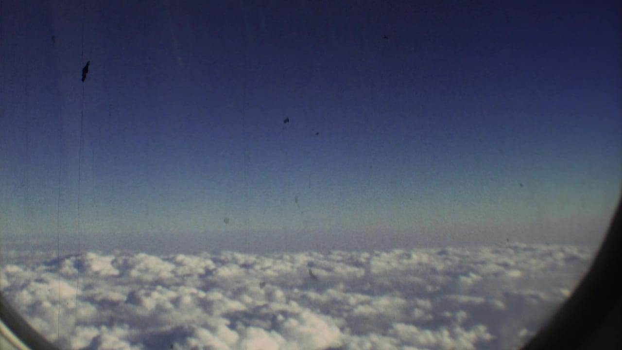 1973 8mm view from an aircraft window, showing cloud tops below a blue sky with visible film grain and window framing.