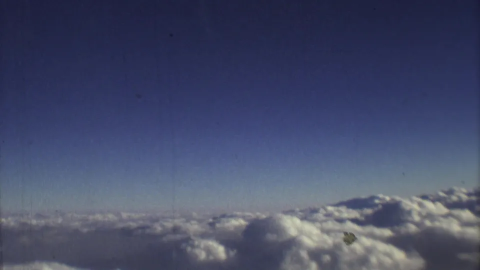 Aerial 8mm frame showing blue sky, distant horizon, and sunlit cloud tops during a 1973 flight near Mexico City.